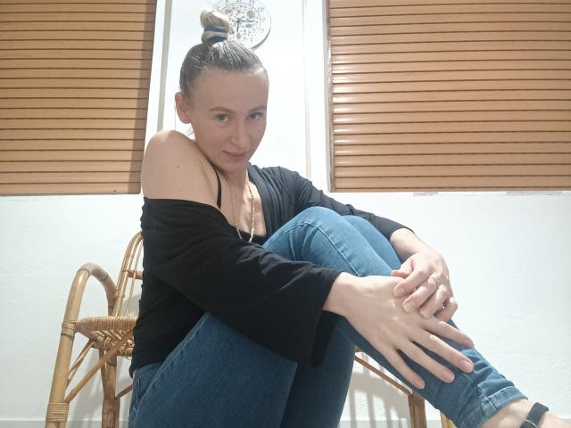Saveta with her tits slightly visible in a black off-shoulder top, sitting on wicker chairs, knees up, hands on her legs, wearing blue jeans, and her hair in a bun.