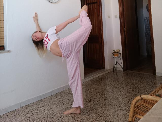 Saveta, blond, zonder schoenen, in roze pyjamabroek, een witte crop top aan, met haar rug tegen de muur, één been hoog opgetrokken, lachend, haar enkel vasthoudend, in een kamer met een granieten vloer..