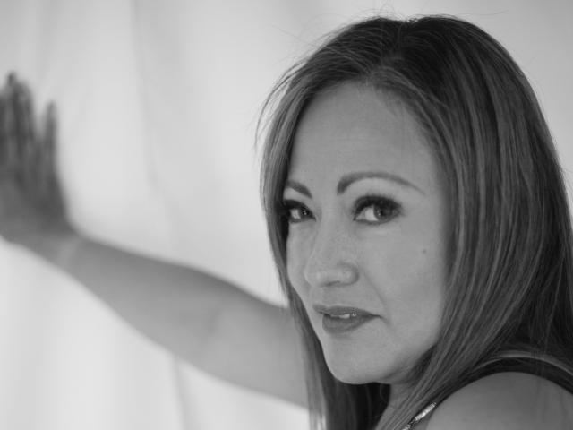 Black-and-white photo of ChristinMoore with straight, medium-length hair, light skin, and subtle makeup, looking over her shoulder with a slight smile, arm raised against a white background.