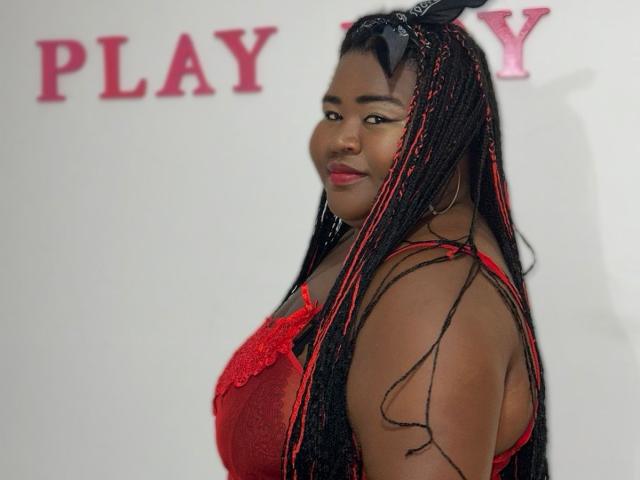 BritannyJhonson with long black and red braided hair, wearing a red lace dress, looking over her shoulder, showing her tits, and smiling. Red lips, hoop earrings, black headband.