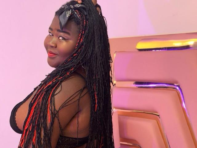 BritannyJhonson with long black and red braided hair, dark skin, red lipstick, black lace top, smiling, looking over shoulder, standing next to pink and yellow machine. Boobs visible.