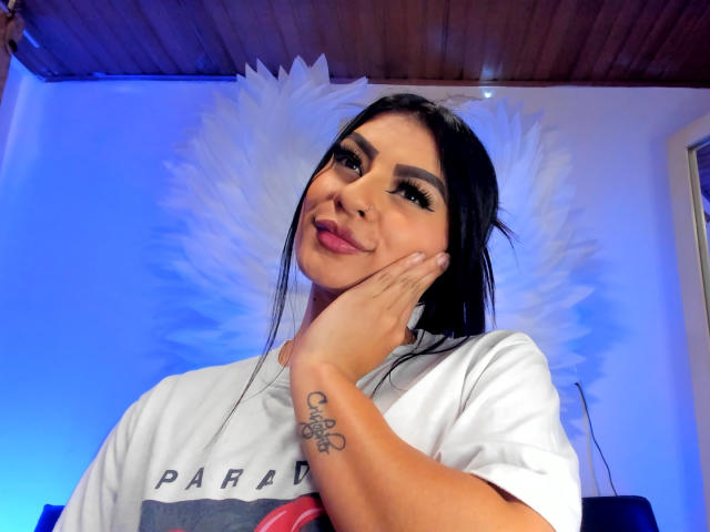 LunaCollinss with dark hair, wearing a white shirt, hand on cheek, smiling, tattoo on wrist, blue-lit background, wooden ceiling, white feather wall art.