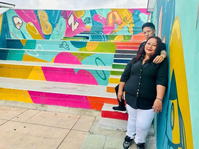 AliceAndTheo, uma mulher grávida com batom vermelho, camisa preta, calças brancas e sapatos pretos, está ao lado de um homem que a abraça, em frente a uma parede com grafites coloridos..