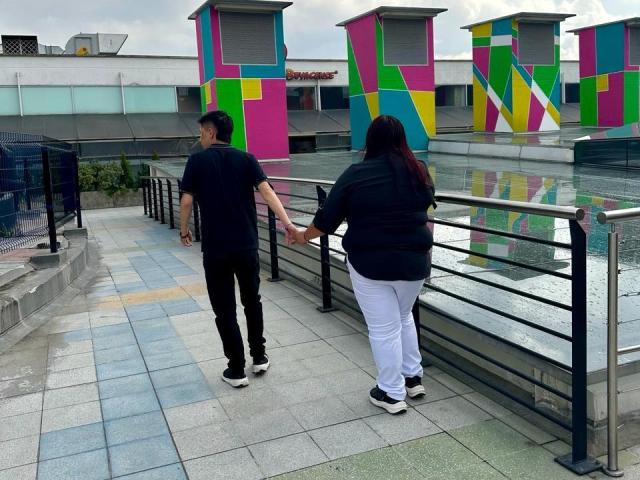 Alice e Theo de mãos dadas, camisa preta, calças brancas, sapatos pretos; Theo, camisa preta, calças pretas, sapatos pretos, caminhando em um telhado de azulejos, colunas coloridas ao fundo..