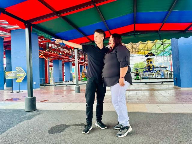 AliceAndTheo, uma mulher corpulenta vestida com uma blusa preta e calças brancas, está ao lado de um homem magro vestido de preto, apontando para baixo de um telhado colorido, ambos sorrindo..