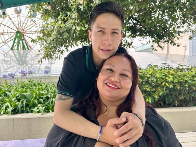 AliceAndTheo, uma mulher asiática corpulenta com cabelo ruivo, está sentada em frente a Theo, um homem asiático magro com cabelo preto, que a abraça por trás, sorrindo, ao ar livre..