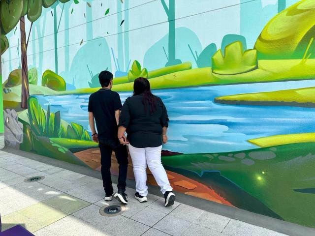 Alice e Theo em pé, com os traseiros próximos, camisa preta, calças pretas, sapatos pretos; outra Alice e Theo, camisa preta, calças brancas, sapatos pretos, de frente para um mural colorido de colinas verdes e água azul..