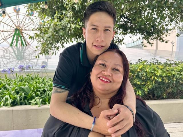 AliceAndTheo com cabelo preto curto, pequena tatuagem no braço direito, camisa preta, abraçando AliceAndTheo com cabelo ruivo, camisa preta, sorrindo, mãos no colo, ao ar livre em meio à vegetação..