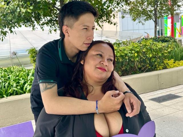 AliceAndTheo, a chubby woman with big boobs in a black jacket and red bra, sits while Theo, a slim man with tattoos, hugs her from behind.