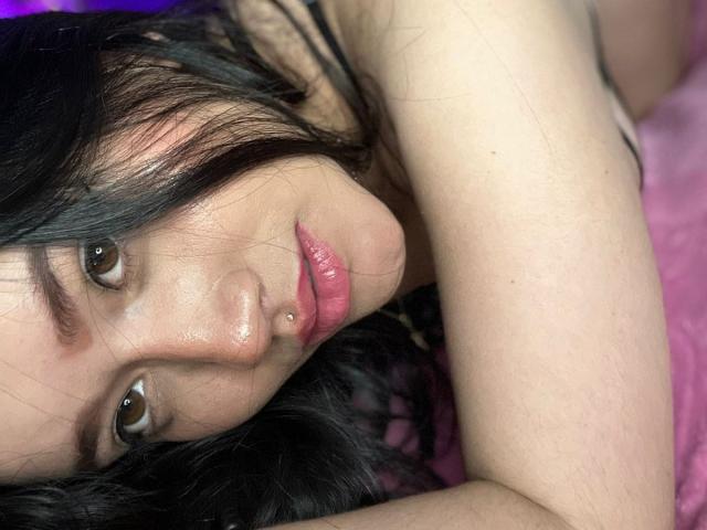 VeronicaWins has a cute face, dark hair, and a small nose piercing. Her pink lips are slightly parted, and she's lying on a pink surface with a relaxed expression.