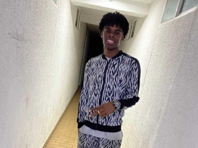 ElroyBlack with dark skin, short curly hair, wearing a black and white zebra-patterned shirt and pants, standing in a hallway, smiling, wearing a wristwatch.