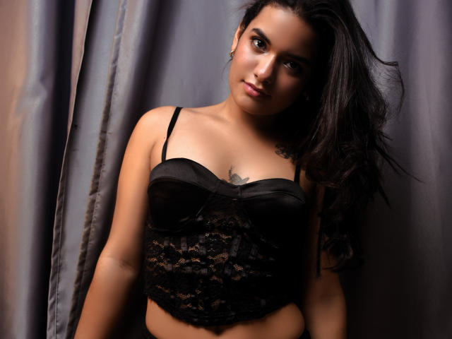 AbbyZalazar with dark lace bra, medium tits, black hair, slight smile, tattoo on chest, brown eyes, standing against gray curtain, looking at camera, sexy, confident, attractive.