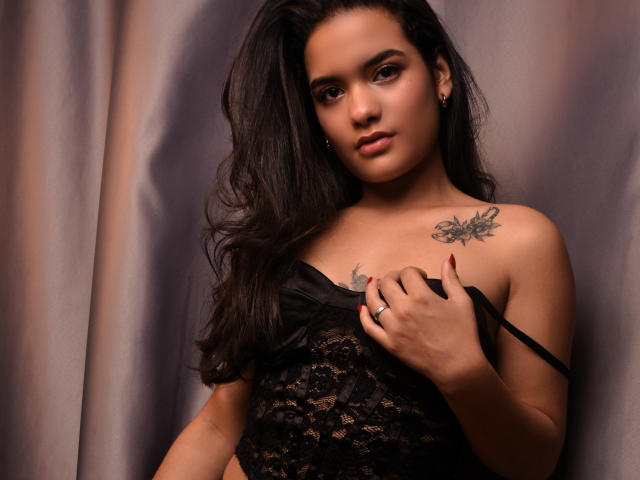 AbbyZalazar with long dark hair, lace black top, holding strap, showing tattoo on right shoulder, ring on finger, and red nails, with a confident, slightly sultry expression.