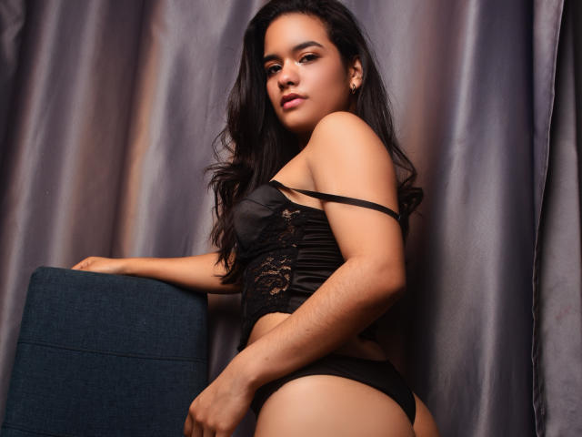 Abby Zalazar, con cabello largo y negro, vestida con un corpiño negro con cordones y ropa interior negra, apoyada en una silla azul, mostrando su trasero y un poco de busto, mirando a la cámara..