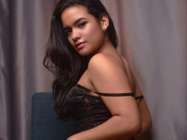 AbbyZalazar with long dark hair, wearing a black lace strap dress, sitting on a blue chair, looking over her shoulder, showing her bare shoulder and a small earring.