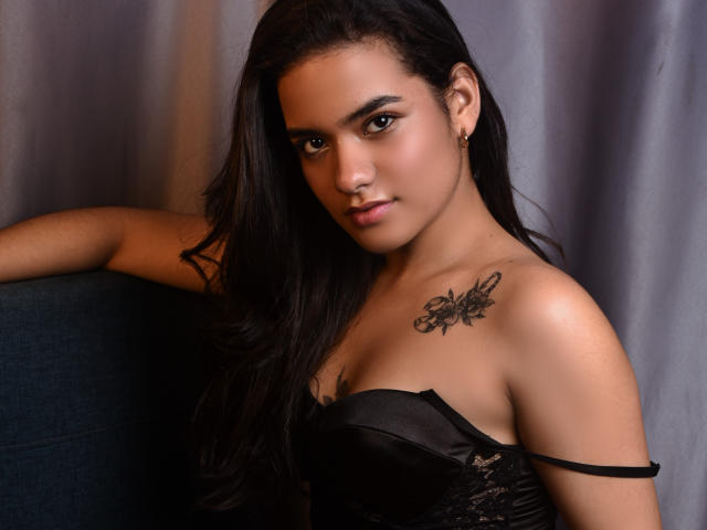 Abby Zalazar, con cabello oscuro, ojos marrones y piel morena, vestida con un corpiño negro con cordones, mostrando un tatuaje de una mariposa en su hombro derecho, apoyada en una silla azul..