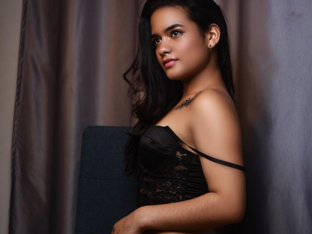 Abby Zalazar, con cabello oscuro, piel marrón media y senos pequeños, vestida con una blusa negra de encaje con un hombro descubierto, está de pie junto a una silla azul, mirando a la cámara..
