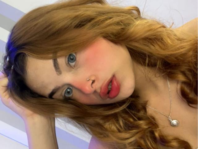 ShantalSummer has wavy brown hair, blue eyes, pink blush, nose ring, red lips, and a silver necklace with a small pendant. Her hand's in her hair, looking at camera.