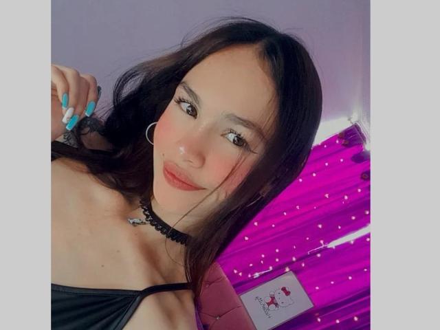 LiliiMoore with dark brown hair, blue nails, black choker, red lips, and hoop earrings, showing her black strap top and small tits, smiling at the camera.