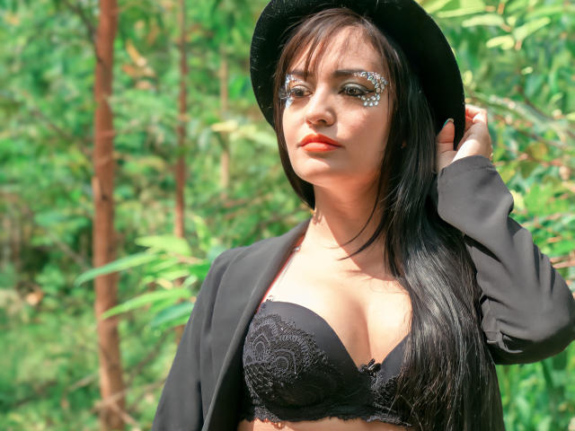 SilvannaReyes with long black hair, wearing a black hat, black lace bra showing her tits, and a black jacket, standing in a green forest, with glittery eye makeup.
