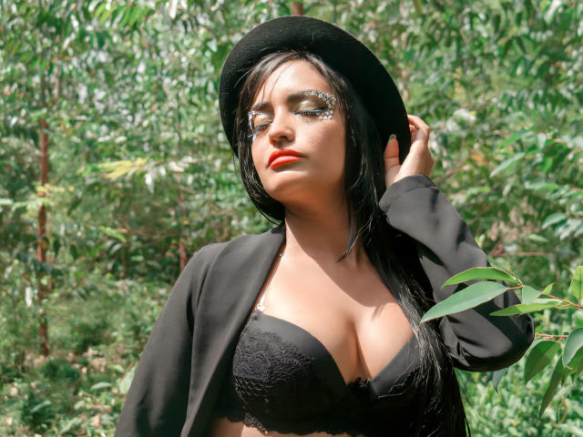 SilvannaReyes with big tits in a black lace bra, wearing a black hat and jacket, standing in a forest, touching her hat with green leaves in the background.