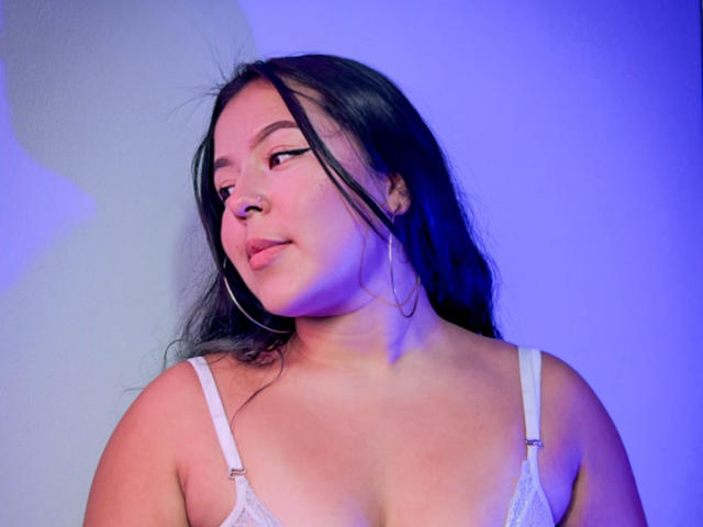 BarbaraHoneyy with long black hair, wearing white lace bra, looking sideways with a slight smile, large hoop earrings, blue-purple light on her face, and soft pink skin.