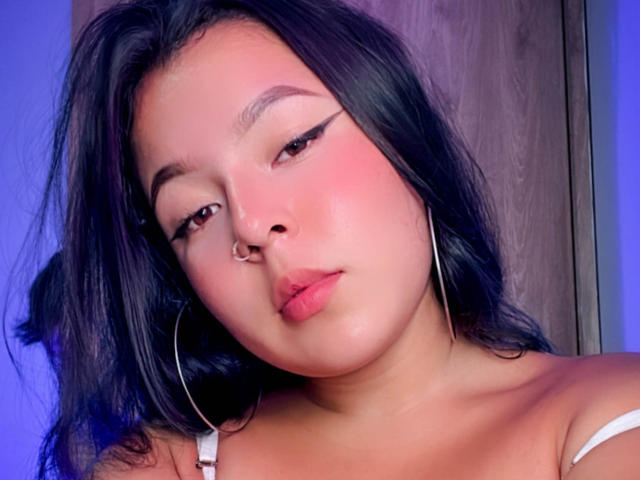 BarbaraHoneyy has black wavy hair, a nose ring, and wears hoop earrings with pink blush on her cheeks. She's looking slightly to the side with pink lips.