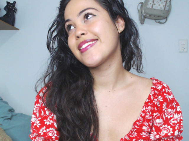 SharonBurtton with wavy black hair, red floral dress, pink lipstick, smiling, looking up, light skin, small tits, soft pink nipples, cute, white wall, shelf, cat figurine in background.