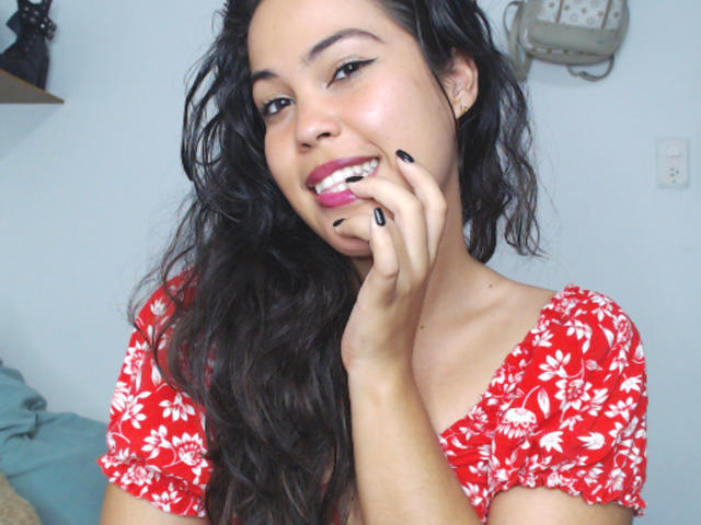 SharonBurtton with long black hair, red floral dress, smiling, pink lipstick, black nail polish, touching her lip with her hand, looking cute, light skin, bedroom setting.
