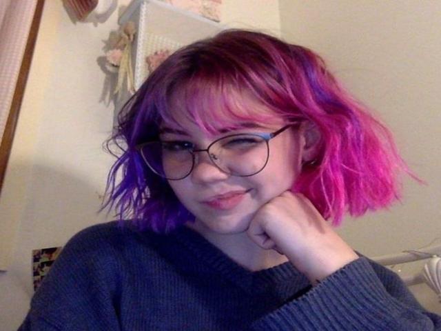 LeilaAngel with pink and purple hair, glasses, and a grey sweater, resting her chin on her hand, smiling slightly. Her ass is not visible.