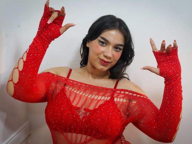 Milly Johnson, met donker, golvend haar, een rode, glinsterende top met een open schouder, en bijpassende rode mouwloze armwarmers, lachend, met de handen omhoog, waarbij een rode bh onder de top zichtbaar is, met een gemiddelde borstgrootte, en een lichte huid..