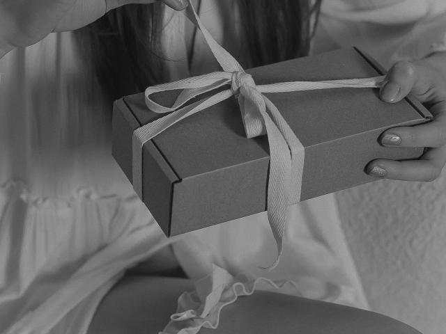 JaneRoller's hands holding a gift box with a white ribbon, wearing a white dress, visible thigh, and nails with shiny polish. Black and white image.