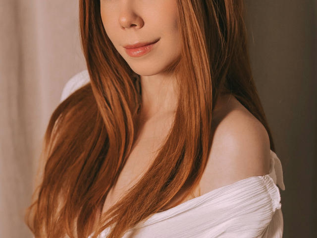 JaneRoller with long, straight, red-brown hair, off-shoulder white top, showing bare shoulders, subtle smile, soft light, smooth skin, gentle expression, slight blush on cheeks.