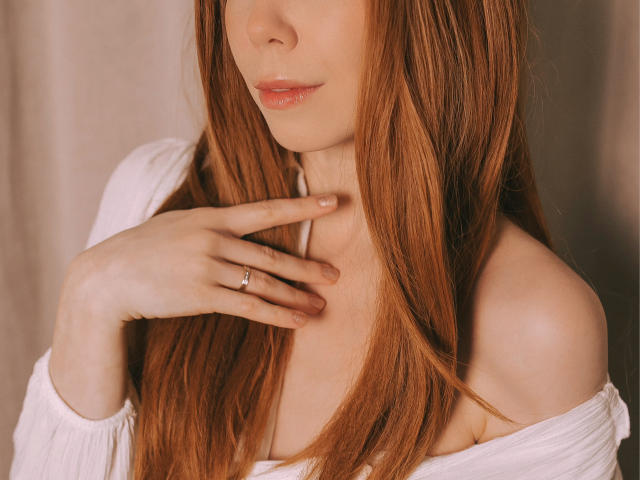 JaneRoller has long red hair, pale skin, and a subtle smile. She's touching her neck with her right hand, wearing a white off-shoulder top. She's wearing a simple ring.
