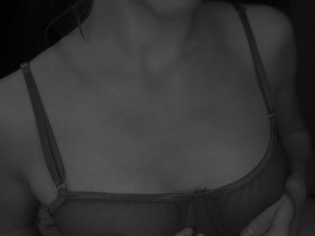JaneRoller's tits in a dark, sheer bra, visible cleavage, and a hand slightly pulling up the cup. Black-and-white, soft lighting, gentle shadows.