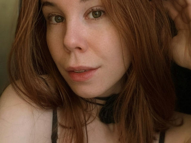 JaneRoller has light green eyes, reddish-brown hair, and freckles on her face. She's wearing a black choker with a ribbon and a black spaghetti strap.