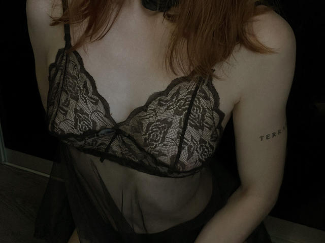 JaneRoller in black lace lingerie, showing her tits in a sheer top, with a "TERA" tattoo on her right arm, wearing a black choker, and with red hair.
