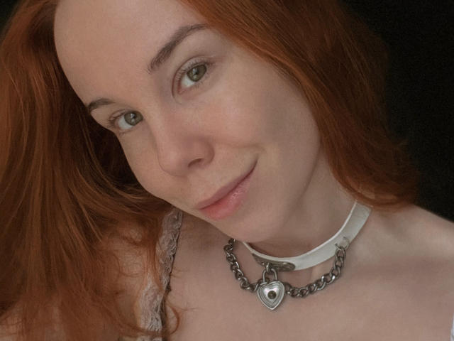 JaneRoller has red hair, green eyes, and a cute smile. She's wearing a white collar with a heart-shaped silver pendant and lace on her shoulder.
