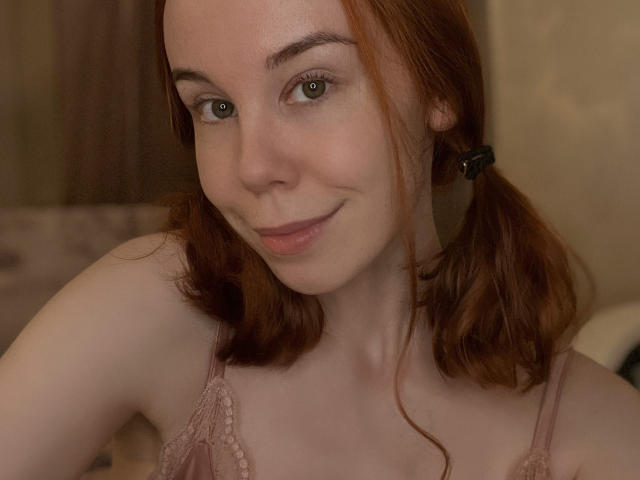 JaneRoller with red pigtails, wearing pink lace lingerie, has fair skin, green eyes, and a slight smile. Her small tits are visible through the lace.