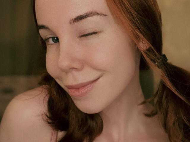 JaneRoller has light skin, winking with a slight smile, brown hair in pigtails, and is showing her bare shoulders. Her face has a cute, playful expression.