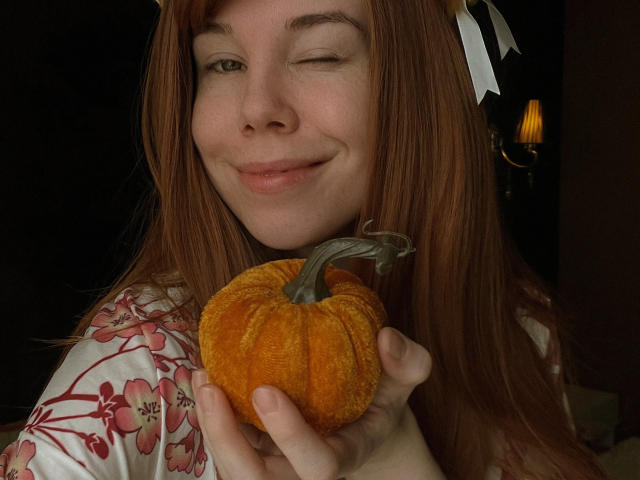 JaneRoller with red hair, winking, holding a small orange pumpkin, wearing a floral shirt, white ribbons in hair, dimly lit room, smiling, cute, pumpkin in hand.