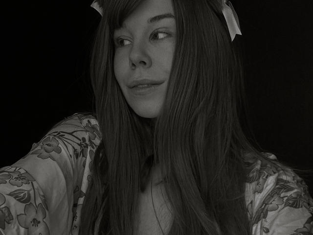 JaneRoller has long, straight brown hair, a floral shirt, and a white ribbon in her hair. She's looking to the side with a slight smile. Black and white photo.