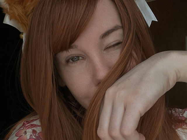 JaneRoller with red hair, winking, and covering her mouth with her hand, showing her pale skin and freckles. She's wearing a floral top and a white hairpiece.