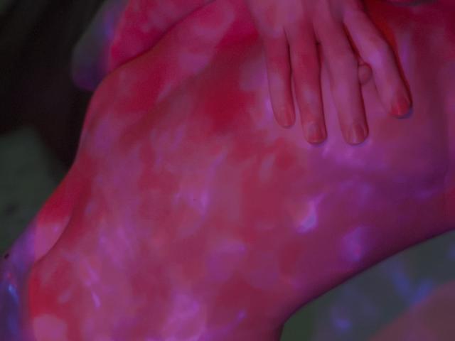 JaneRoller's pink-tinged, smooth belly with a hand resting on it, showing her fingers and slightly curled nails, in a soft, colorful light.