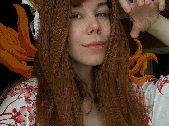 JaneRoller has long red hair, orange fox ears, and a white floral top. She's smirking with green eyes, pale skin, and a hand up. Her tits are small.