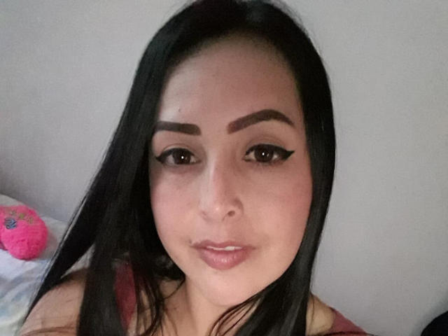 ChristinSanders with long black hair, dark eyebrows, and winged eyeliner, showing off her pouty pink lips and slightly visible nose ring. Pink stuffed toy on the left.