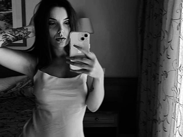 Selfie en escala de grises de JuliCandy con cabello largo y oscuro, aro en la nariz, brazo tatuado, sosteniendo un teléfono, vistiendo una camiseta blanca sin mangas, de pie junto a una ventana con cortinas florales. Se ven los senos..