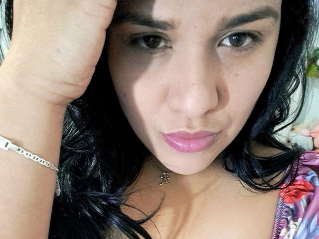 VivianaThomson has dark hair, brown eyes, pink lipstick, and a silver bracelet on her right wrist. She's slightly pouting with her hand near her head.