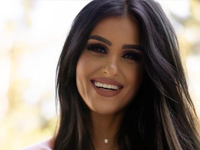 SelenaCruz has long black hair, dark eyebrows, and is smiling with white teeth, wearing makeup and a pink top, with a small pearl necklace.