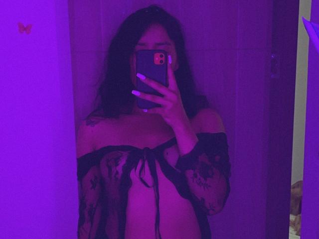 ElizabethMacy with long dark hair, wearing a sheer black lace robe, exposing her tits, taking a mirror selfie with purple neon lighting, holding a phone with white nails.